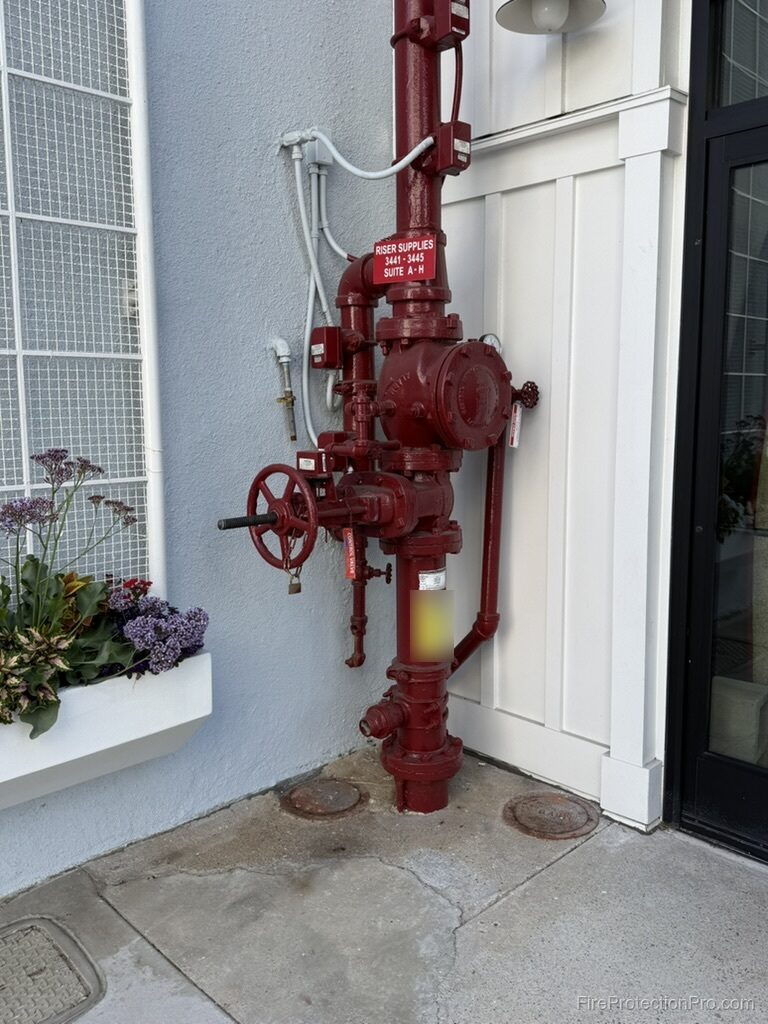 Exterior wall-mounted fire sprinkler riser painted red, with handwheel control valve, supervisory switch conduit, drain piping, and a suite identification placard