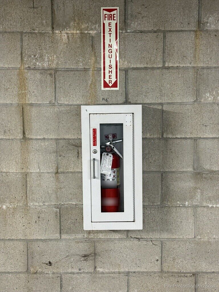 Semi-recessed white fire extinguisher cabinet on a concrete block wall with a vertical FIRE EXTINGUISHER arrow sign mounted above it