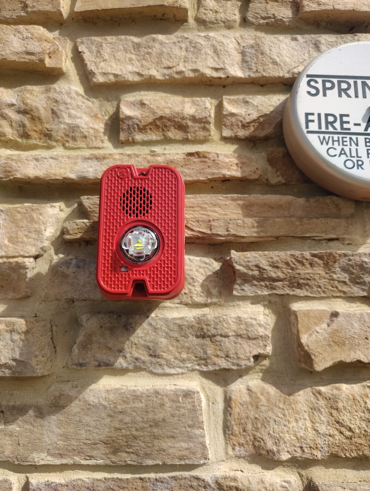 Wall-mounted horn/strobe notification appliance next to a sprinkler fire alarm sign on a commercial building exterior