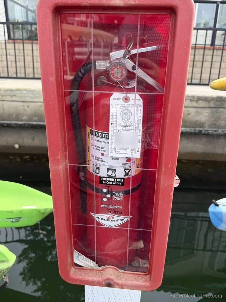 Weathered red outdoor fire extinguisher cabinet with a break-panel front, mounted at a waterfront dock